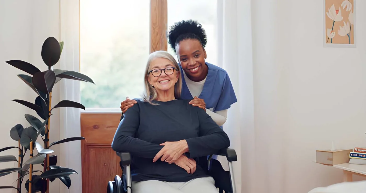 An older woman who uses a wheelchair and her caregiver smiling together at home